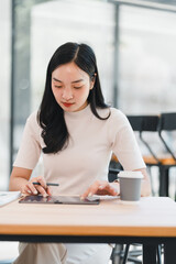 Young Asian woman using digital tablet with stylus in modern office, focused and working, coffee...