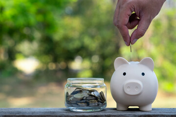 A hand drops a coin into a piggy bank beside a jar of coins, symbolizing savings and financial...