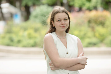 young woman wearing a white dress and a pearl necklace is standing in a park. She is looking at the...