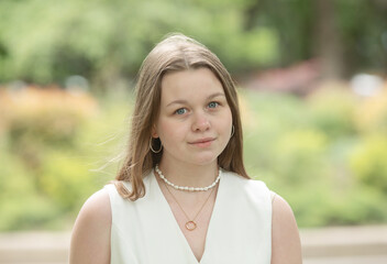 young woman wearing a white dress and a pearl necklace is standing in a park. She is looking at the...