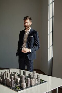 A young man stands in a professional suit, gesturing toward a detailed architectural model. The focus is on urban development.