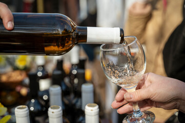 Close-up of a person's hand pouring white wine into a glass, many bottles of wine nearby