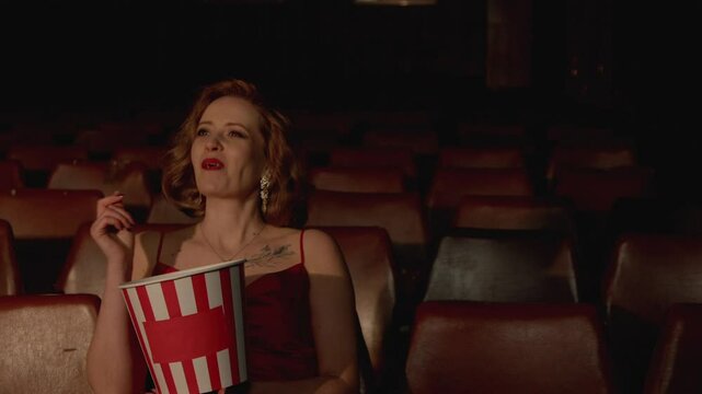 Cheerful pretty middle aged woman in stylish dress enjoying leisure, having fun, watching comedy movie and eating popcorn in empty retro style cinema.