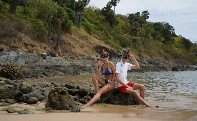Fototapeta premium A young couple wear goggles and snorkels siting on a rock by the water