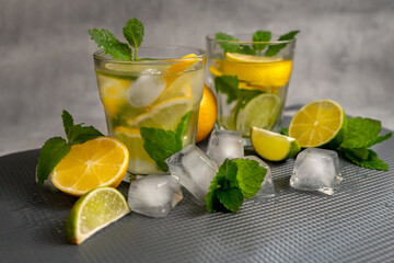 Two glasses of lemonade drink with mint and lemon on a gray textured background with scattered ice, mint and citrus fruits. Food photo of summer citrus lemonade.