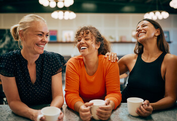 Connection over coffee. Three laughing women of varied backgrounds share laughter during a break. The scene radiates joy, friendship, and trust in a cozy cafe.
