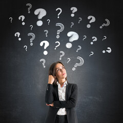 Thoughtful businesswoman standing near black wall with many hand-drawn question marks floating above her head, representing curiosity and problem solving.