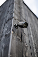 A sleek black security camera mounted on the corner of a modern black wooden building, angled for wide outdoor surveillance