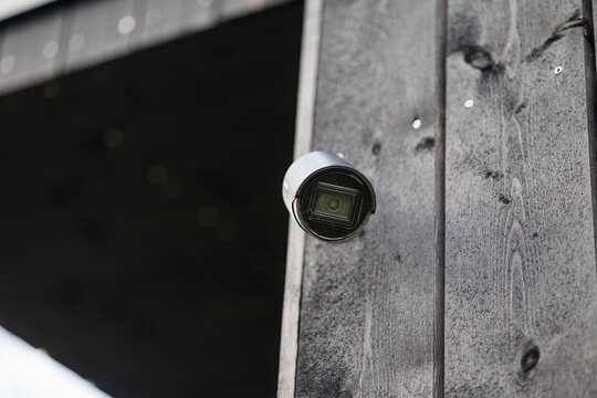 A sleek black security camera mounted on the corner of a modern black wooden building, angled for wide outdoor surveillance