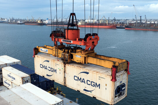 Tauranga, New Zealand 04 30 2025: White containers from shipping company CMA CGM is hanging from spreader of gantry crane in port of Tauranga, new Zealand. In background are general cargo vessels.
