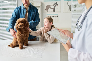 Caucasian girl and her father cuddling their cute dog sitting on medical table in front of them while waiting for check-up