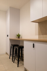 Minimalist kitchen corner with long wood countertop, white cabinets and two black bar stools with a plant decor