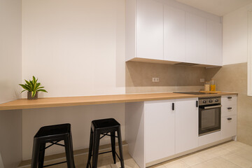 Kitchen interior with long wooden countertop, white cabinetry, black stools and built-in oven in...