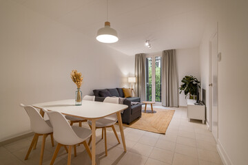 Minimalist dining and living area with white dining table, chairs, gray sofa, and window with natural light