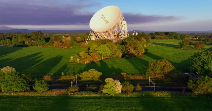 Aerial video of a green countryside landscape with a white radio telescope and a train travelling across in Goostrey - United Kingdom. 