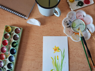 Watercolor drawing, brushes, paints, cup with water on a wooden table. Painter equipment and drawn picture on deck top view