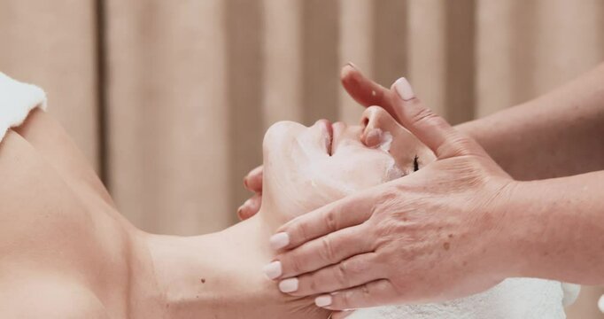 Close-up of a beautician applying moisturizing cream or scrub on a young woman's face in a modern spa salon. The concept of SPA, cosmetology and facial skin care.