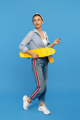 Positive young woman with yellow penny or skateboard posing over blue background