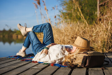 Woman relaxing and napping on pier at lake, face covered by straw hat. Peaceful solitude moment, digital detox and mindful slow living