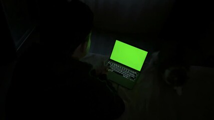 Person sitting on bed watching laptop with green screen for customizable video content.