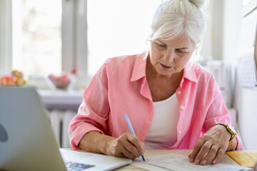 Senior woman working with laptop and documents at kitchen table at her home
