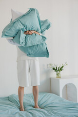 A person is currently standing on a bed while holding a fluffy pillow high above their head, creating a playful atmosphere in the room