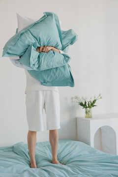 A person is currently standing on a bed while holding a fluffy pillow high above their head, creating a playful atmosphere in the room