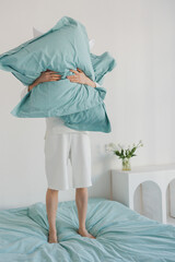 A person is holding a large, soft pillow high above their head while standing on a cozy bed that is covered with colorful linens