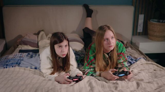 Two teenage girls lying on a bed, playing video games together with relaxed and focused expressions.