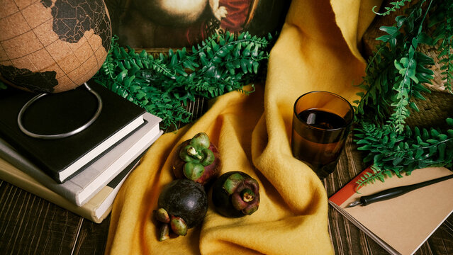 Still life set up on wooden table with three purple Mangosteen fruit, classic painting replica, stack of books, fake green vines, dip pen, notebook, yellow cloth, cork globe, and glass with water.