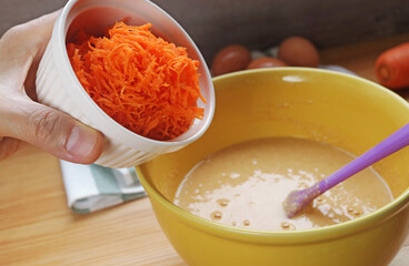 Hand adding shredded carrot into the mixing bowl of batter for baking cake