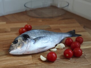 Freshly caught fish displayed with vibrant cherry tomatoes and garlic on a wooden cutting board in a bright kitchen setting