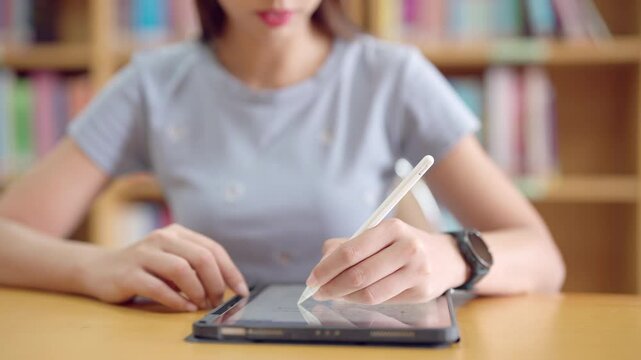 A woman is writing on a tablet with a pen. She is sitting at a table with a stack of books behind her
