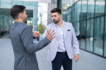 Two businessmen arguing outdoors, one pointing a finger while the other gestures defensively, illustrating a conflict or disagreement in a professional setting.

