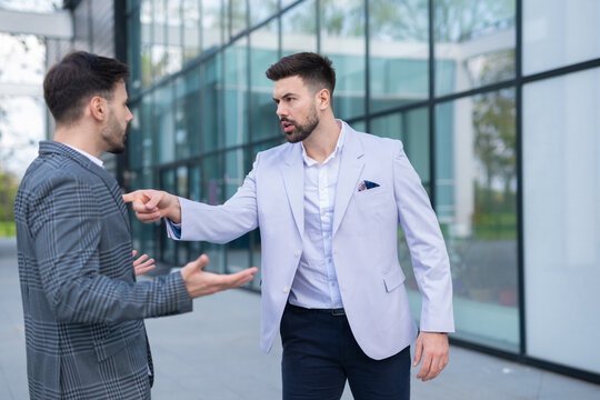 Two businessmen in a heated argument outdoors, one pointing his finger aggressively while the other responds with open hands, showcasing workplace conflict and tension.