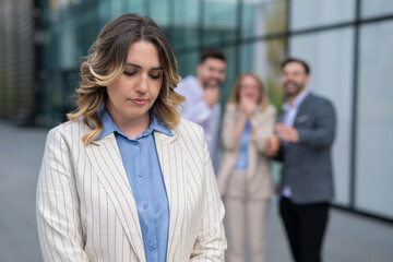 Sad businesswoman facing bullying at work while coworkers laugh behind her