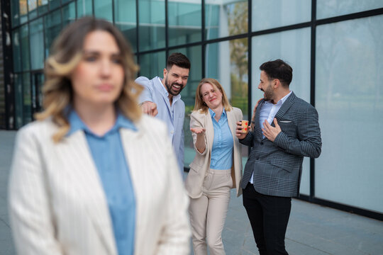 Sad businesswoman standing while colleagues laugh and gossip behind her