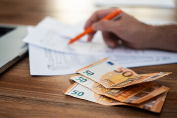 Male freelancer engages in filling tax forms process carefully examining strict requirements and deadlines at workplace with euro banknotes and laptop