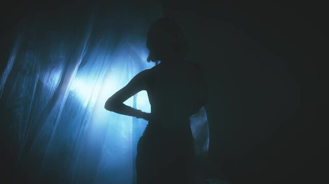 dark silhouette woman walking to neon blue light in night room full smoke fog silk curtains fly rain lightning wind blowing slow motion. Gothic Girl fashion model in sexy black dress. back rear view