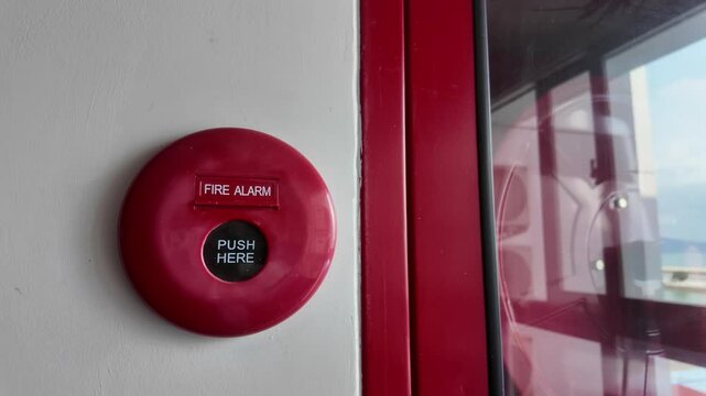Slow motion footage of Red fire alarm on a wall with a PUSH HERE button in the center. It&rsquo;s placed next to a glass cabinet containing fire safety equipment.