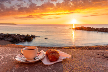 cup of coffee or tea with croissant on a morning embarkment beach with blue sea and beautiful cloudy sunrise or sunset on background, street food breakfast concept