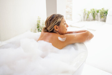 A young woman smiles and takes a bath with fluffy foam. The charming lady has closed her eyes