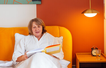 An elderly woman in a white coat reads in bed before going to bed. A beautiful middle-aged lady is...