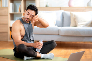Tired young asian man sitting on a yoga mat after a home workout wiping sweat in living room at home, exhausted young man after exercise watching laptop in cozy, self-care and wellbeing.