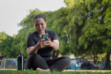 Mindful Asian woman wearing sportswear using smartphone after doing yoga exercise at outdoor public...