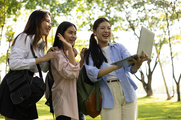 Three young women are standing in a park, smiling and laughing. One of them is holding a laptop, and the others are looking at it. Scene is cheerful and lighthearted
