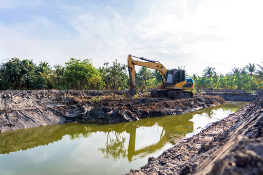 Excavator parked before work in morning sunlight in excavation to expand and lift trench with level area for cultivation of garden furrows with water surrounding them in lowlands central Thailand.