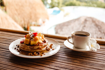 Pancake with mango, fruit, granola and syrup with a cup of hot strong coffee for breakfast. Delicious tropical breakfast on the island at the hotel	