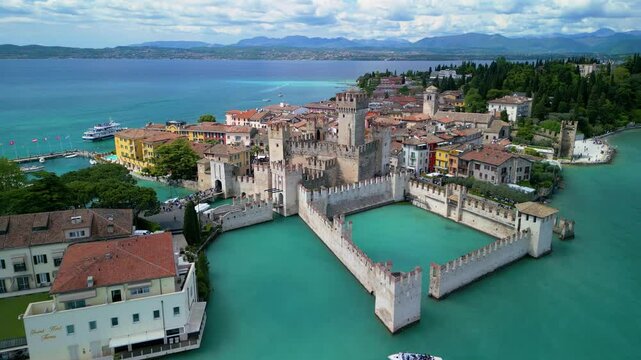 Drone approach to Scaliger Castle in Sirmione, Italy. Historic walls, colorful houses, and turquoise waters of Lake Garda captured on a sunny spring day.