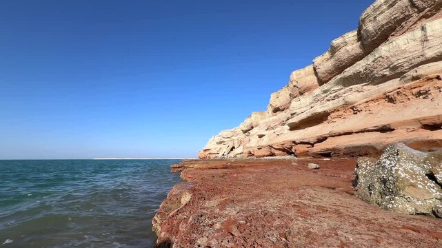 Turquoise waters washing crimson rocky shores of Hormuz Island, exposing unique geological formations beneath clear azure skies in Persian Gulf landscape
